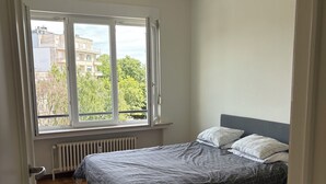 Een slaapkamer met een bed, een radiator, een raam en uitzicht op gebouwen en bomen.
