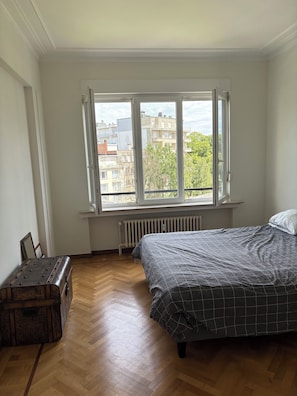 Un dormitorio con un ventanal grande, un radiador, suelo de madera y una cama con colcha estampada.