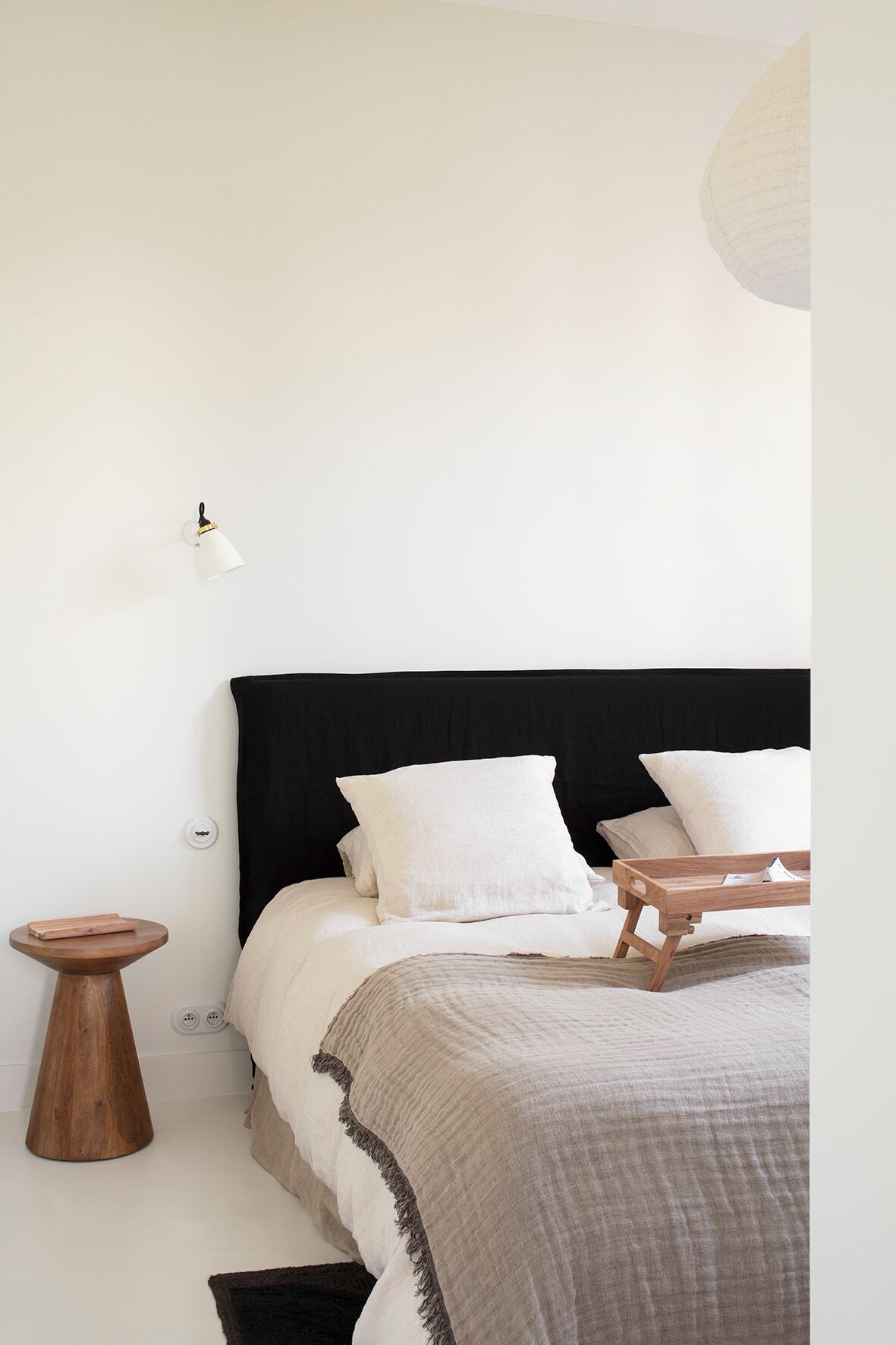 Une chambre à coucher au style minimaliste, avec une tête de lit noire, du linge de lit blanc, une table de chevet et une lampe fixée au mur.