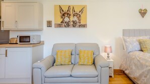 Ein modernes Wohnzimmer mit grauem Sofa, einem kleinen Tisch mit Lampe, einem Bett mit floralem Bettzeug und einer Küchenzeile mit Schränken und einer Mikrowelle.