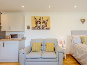 Ein modernes Wohnzimmer mit grauem Sofa, einem kleinen Tisch mit Lampe, einem Bett mit floralem Bettzeug und einer Küchenzeile mit Schränken und einer Mikrowelle.