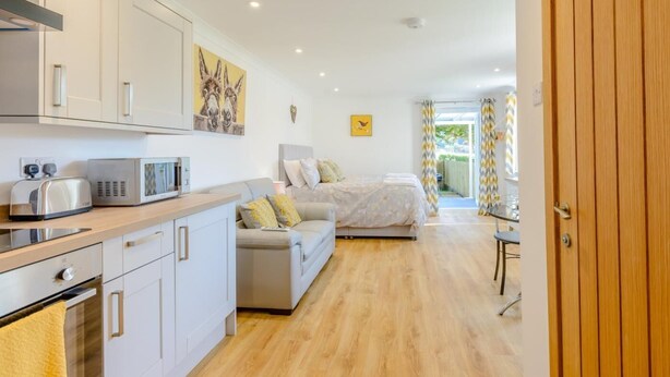 Eine moderne Küche mit weißen Schränken, einer Mikrowelle und einem Toaster. Ein Bett mit grauem Kopfteil und ein Sofa mit gelben Kissen sind ebenfalls vorhanden. Der Raum verfügt über Holzboden und eine Holztür.