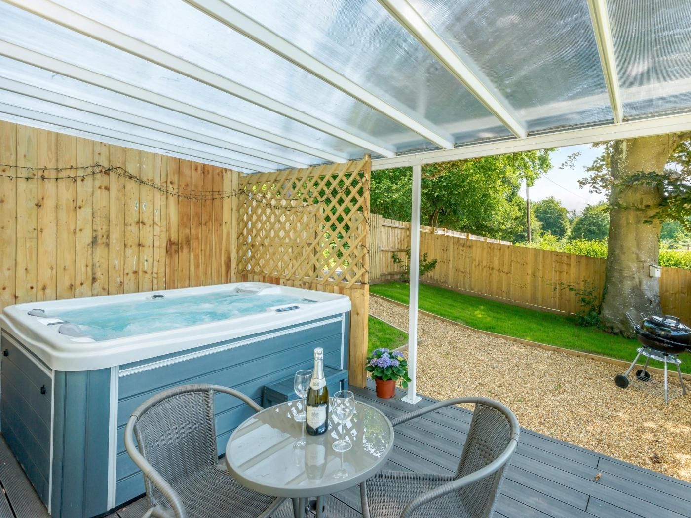A hot tub on a covered deck with wicker chairs and a table, surrounded by a wooden fence and a lawn.