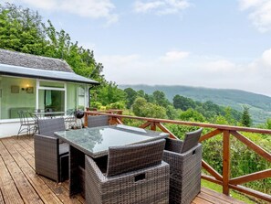 Une terrasse dotée de meubles en osier et d’une table en verre, offrant une vue sur une vallée boisée.