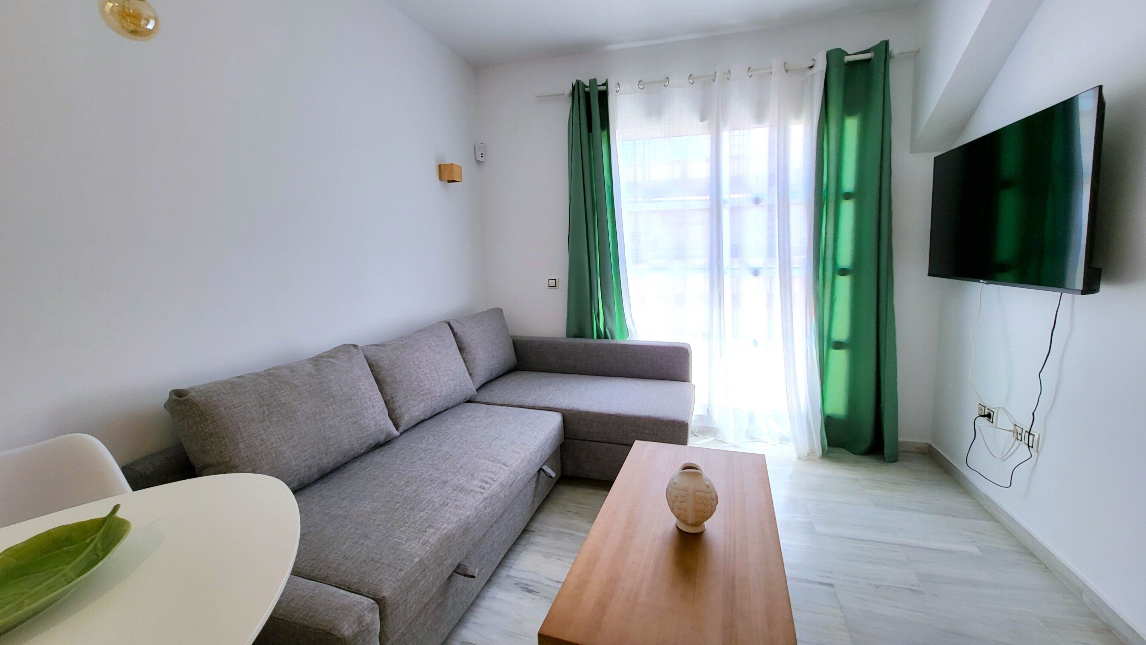 A modern living room with a grey sofa, a wooden coffee table, and a flat-screen TV mounted on the wall.