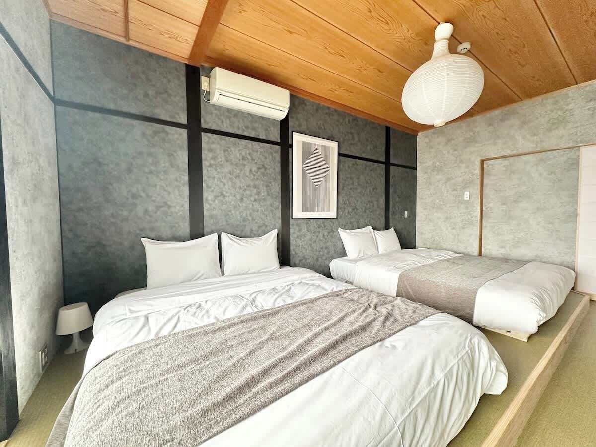A bedroom with a large bed, a smaller bed, and a nightstand. There is a ceiling fan and a wall-mounted air conditioner.