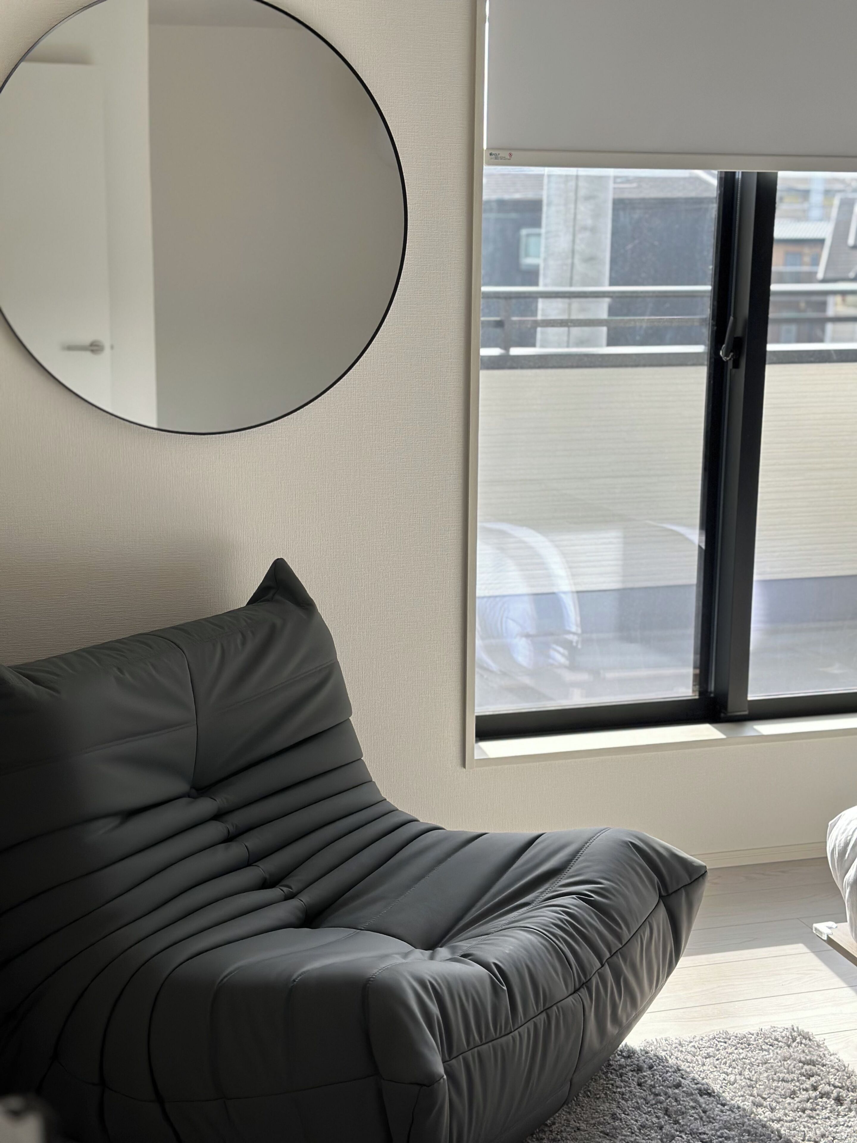 A modern living room with a grey sofa, a round mirror on the wall, and a large window with blinds.