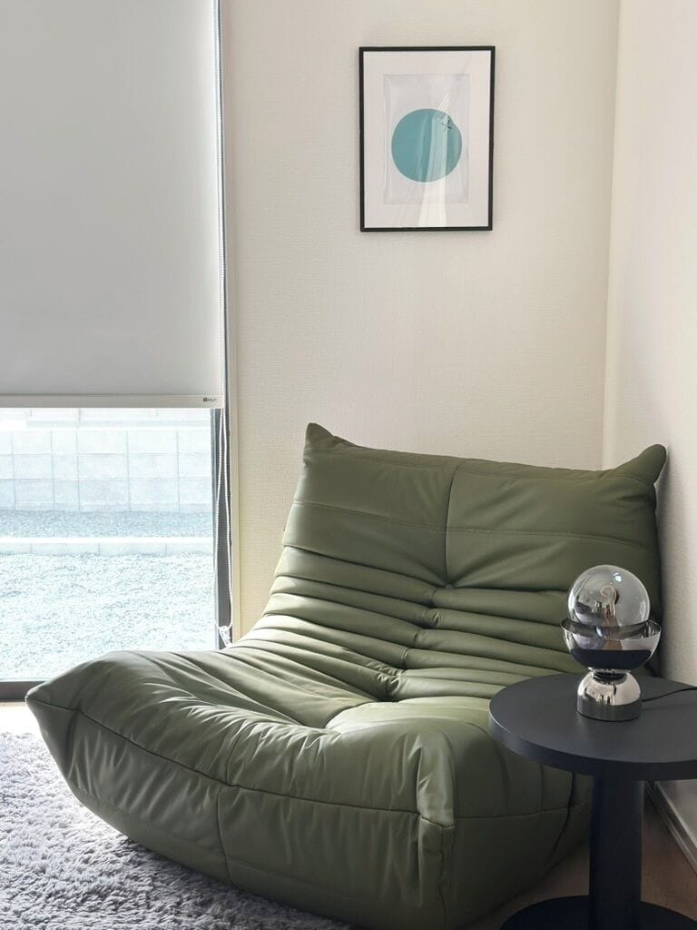 En grön sittpuff i form av en böna, med ett litet bord bredvid, en inramad tavla på väggen och ett fönster med utsikt.