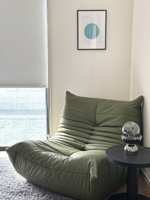 A green bean bag chair with a small table beside it, a framed picture on the wall, and a window with a view. - JP Ozu Units By Restavio (Ozu)
