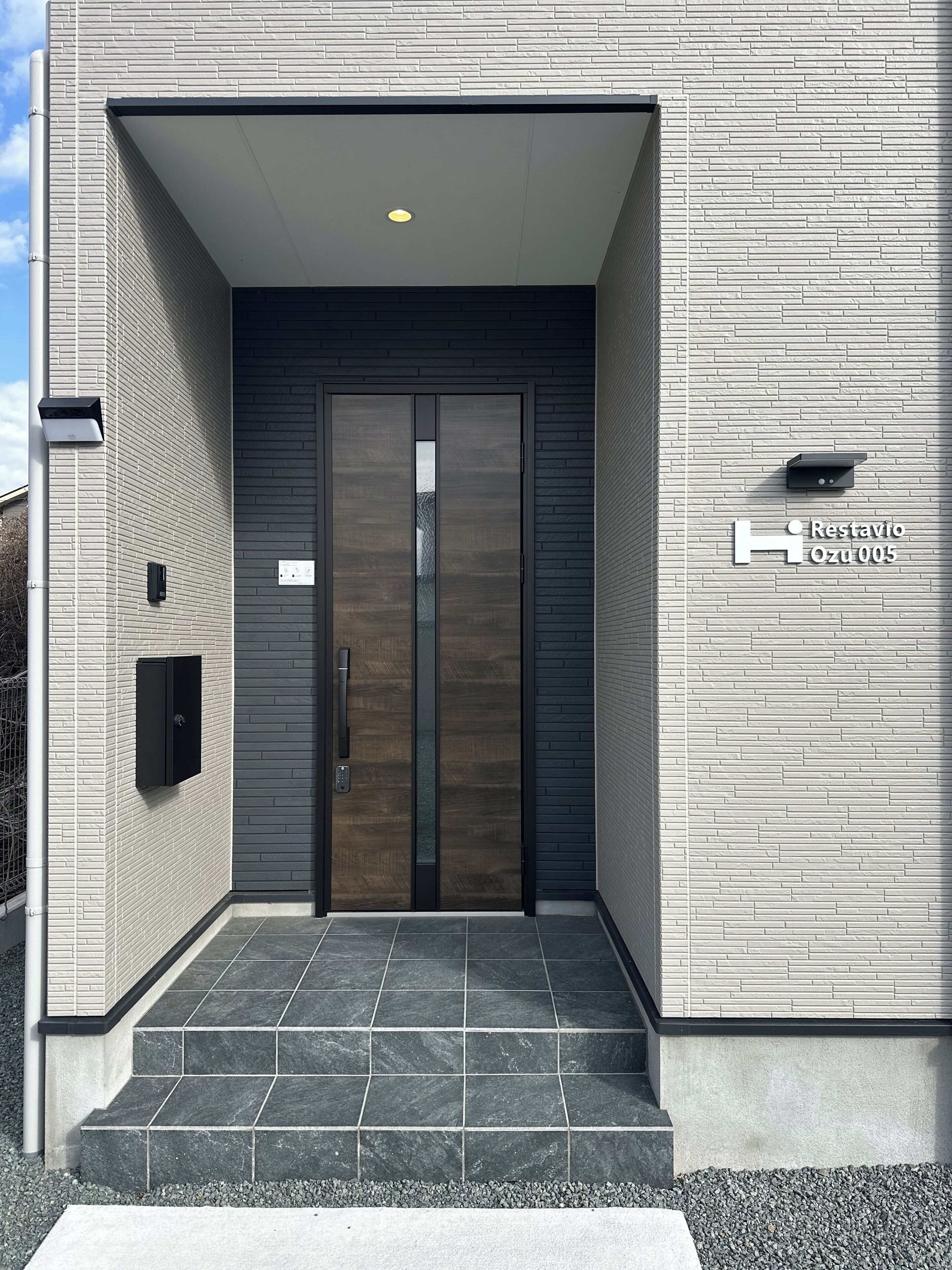 Une entrée moderne avec une porte en bois foncé, une petite boîte aux lettres et une enseigne indiquant « Restavio Ozu 005 ».
