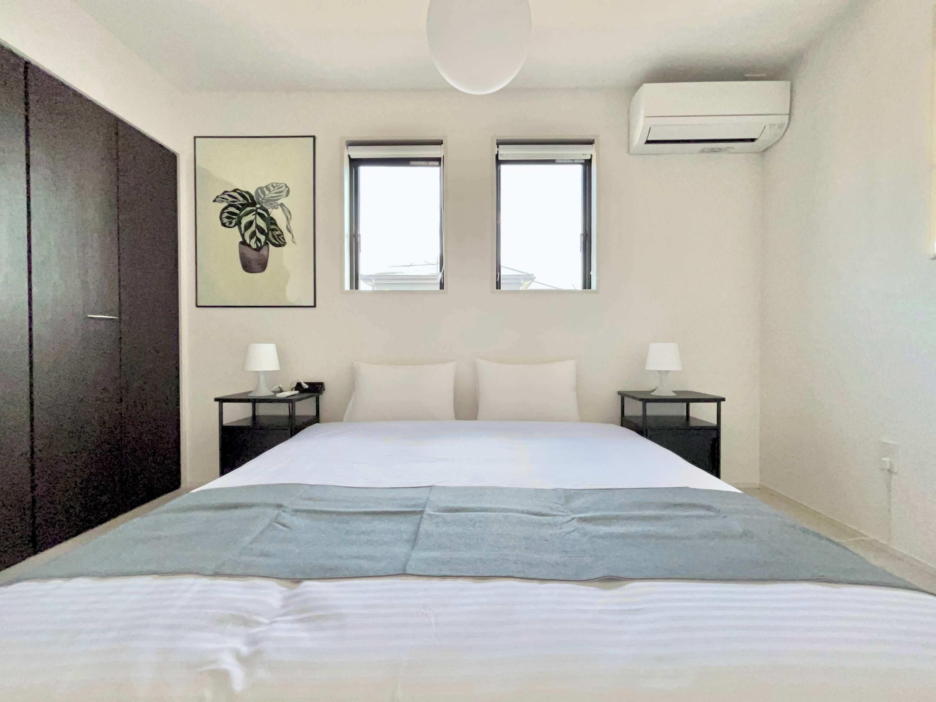 A bedroom with a bed, two bedside tables, a nightstand, a wall-mounted air conditioner, and two windows.