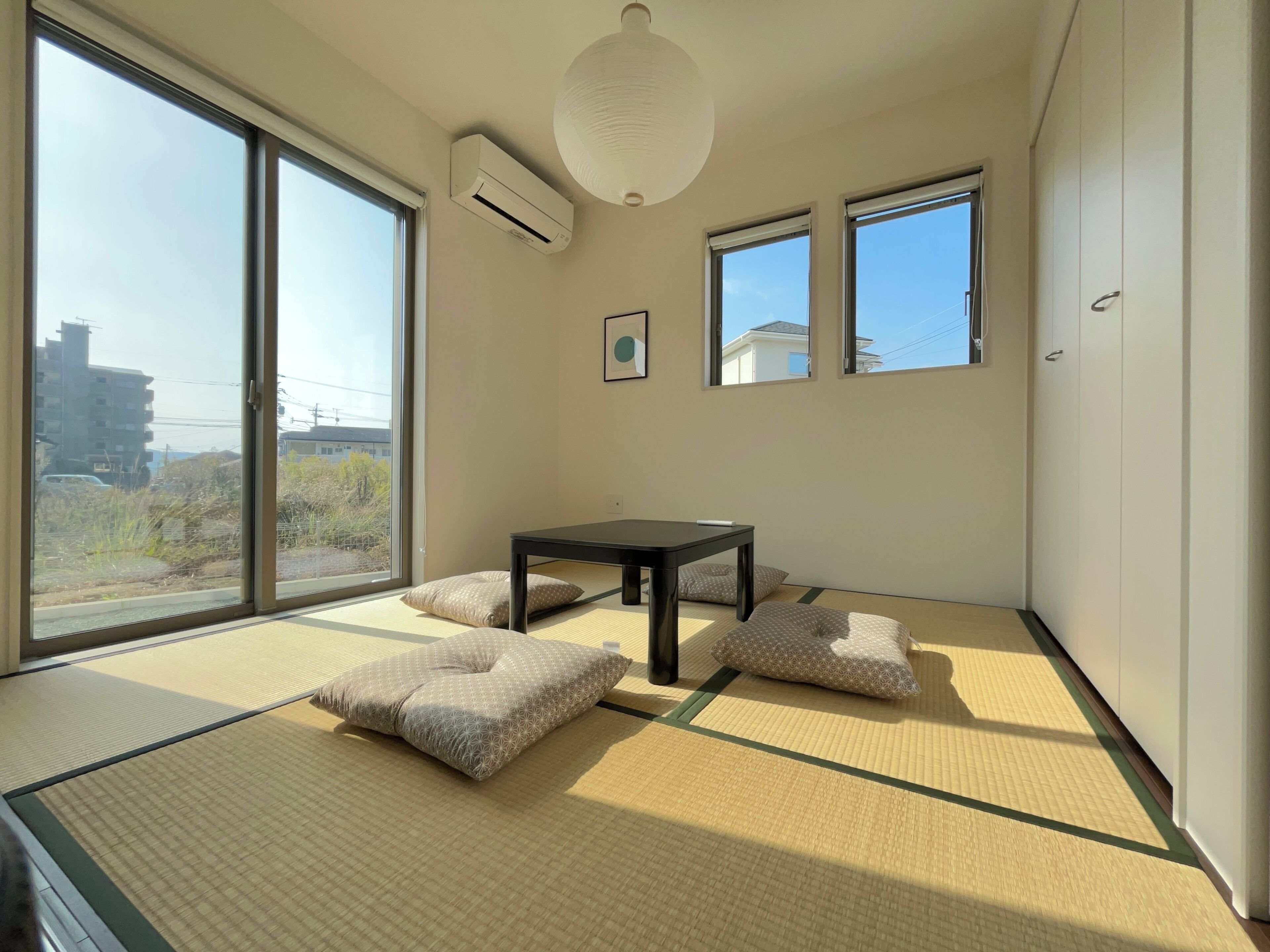 A room with tatami flooring, a table, cushions, and large windows offering a view of buildings and vegetation.