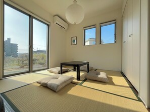 A room with tatami flooring, a table, cushions, and large windows offering a view of buildings and vegetation. - JP Ozu Units By Restavio (Ozu)