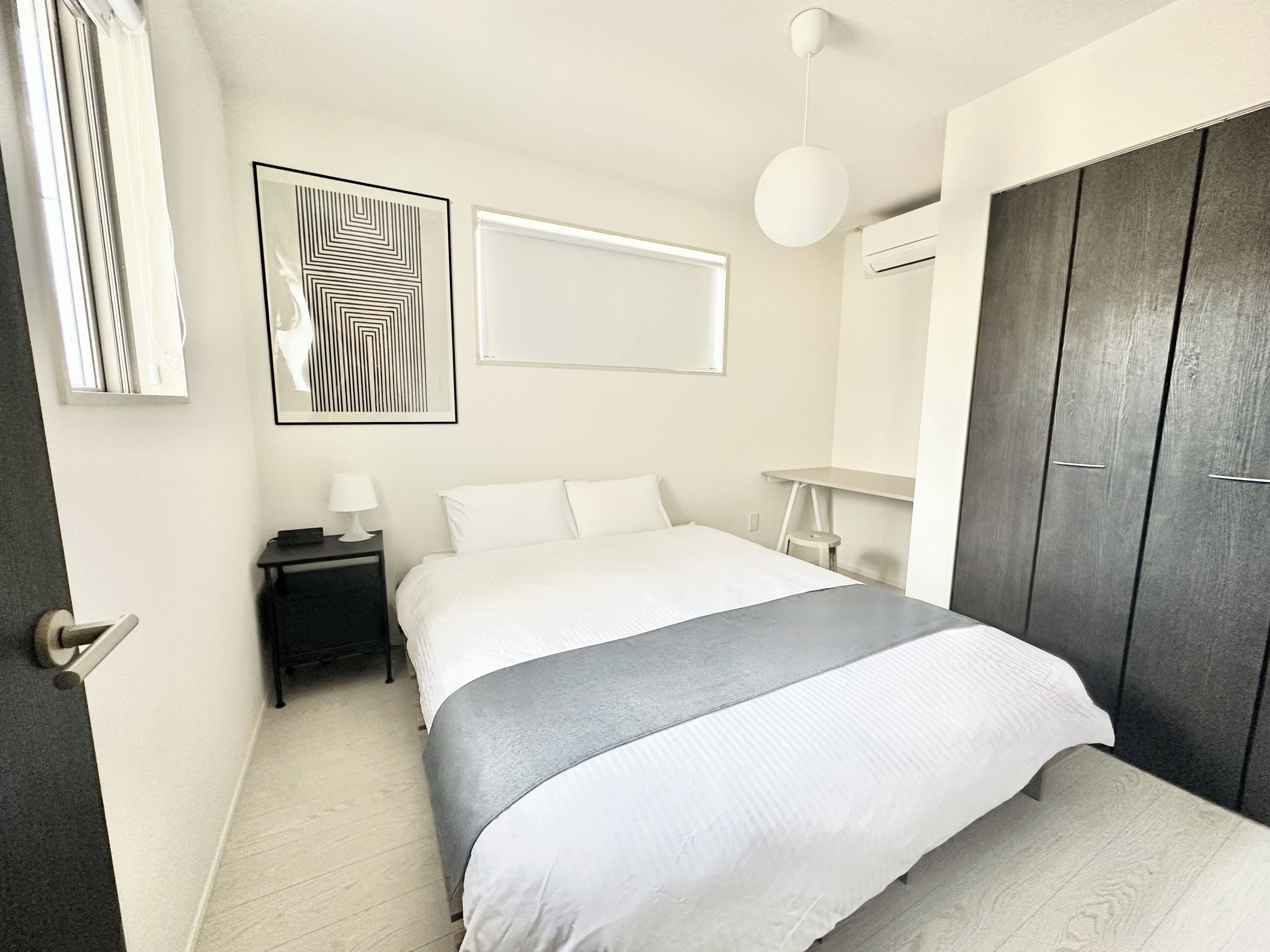 A modern bedroom with a bed, bedside table, lamp, artwork, wardrobe, and a hanging light fixture.