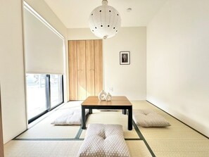 A room with tatami flooring, a wooden door, a table with cushions, and a hanging light fixture. - JP Ozu Units By Restavio (Ozu)