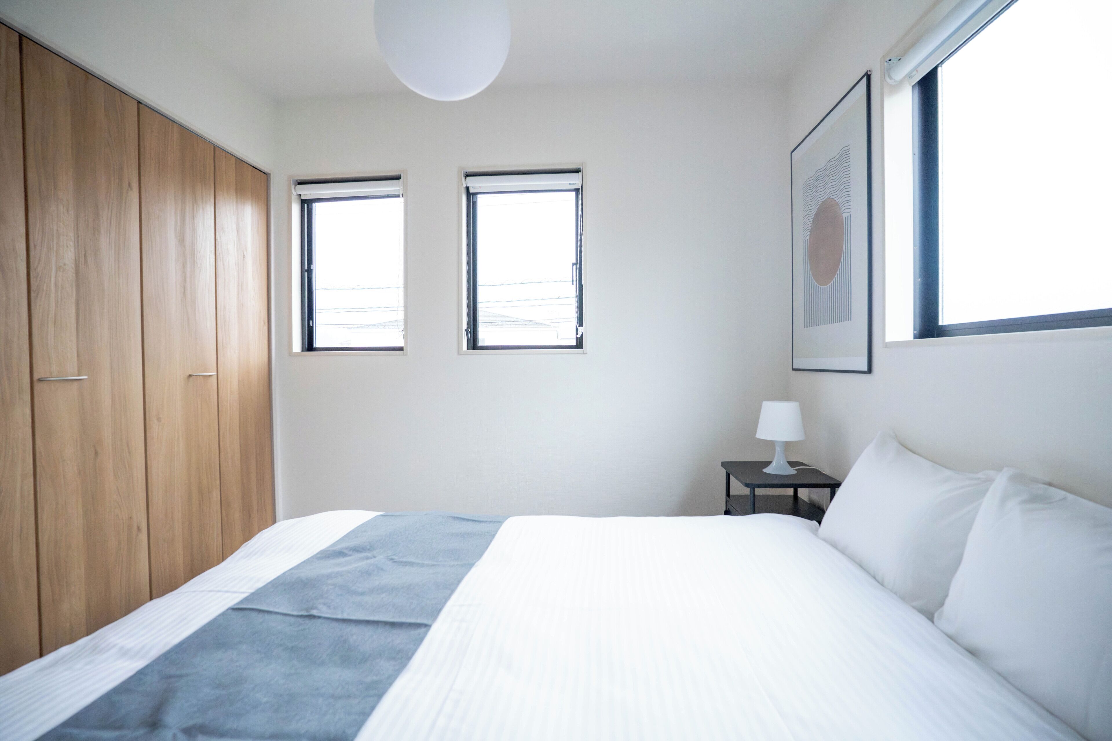 A bedroom with a bed, a nightstand, a lamp, a wardrobe, and two windows.