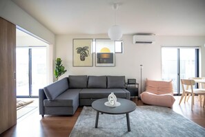 A modern living room with a grey sofa, a round coffee table, and a pink chair. There are two framed artworks on the wall, a potted plant, and a ceiling light fixture.