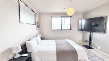 A modern hotel room with a large bed, a flat-screen TV mounted on the wall, a bedside table with a lamp, and a window providing a city view.