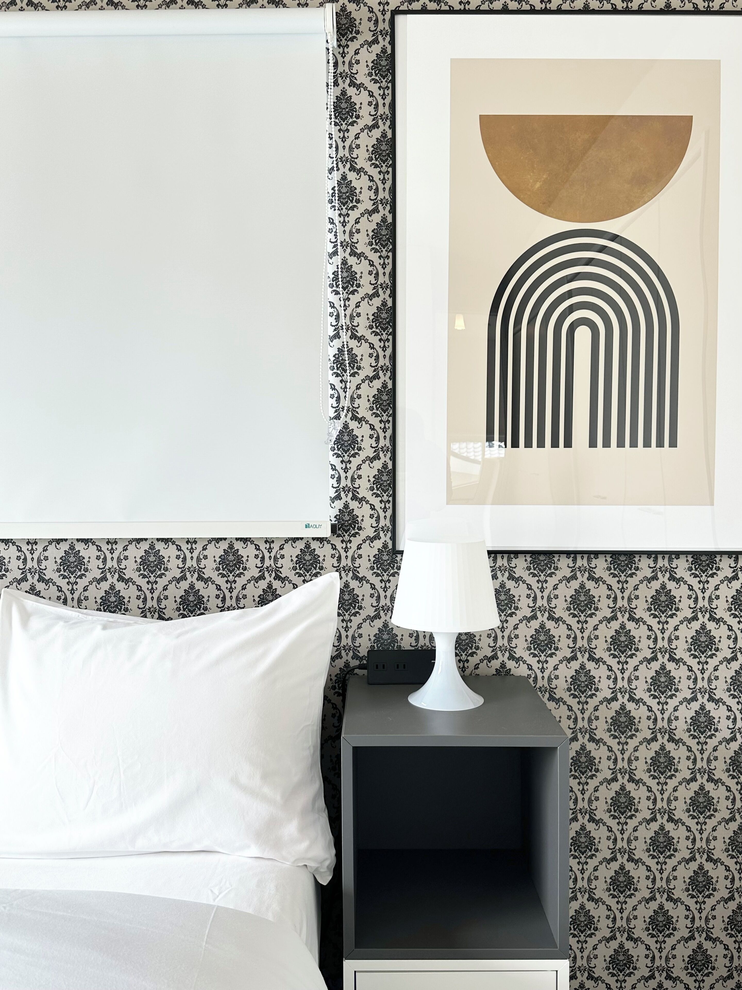 A neatly made bed with white linens, a black nightstand with a white lamp, and a framed abstract artwork on the wall.