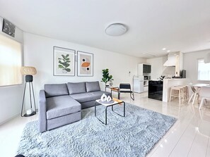 Ein modernes Wohnzimmer mit einem grauen Sofa, einem Couchtisch und einem blauen Teppich. Im Hintergrund befindet sich eine Küche mit weißen Schränken sowie ein Essbereich mit Tisch und Stühlen in Weiß.
