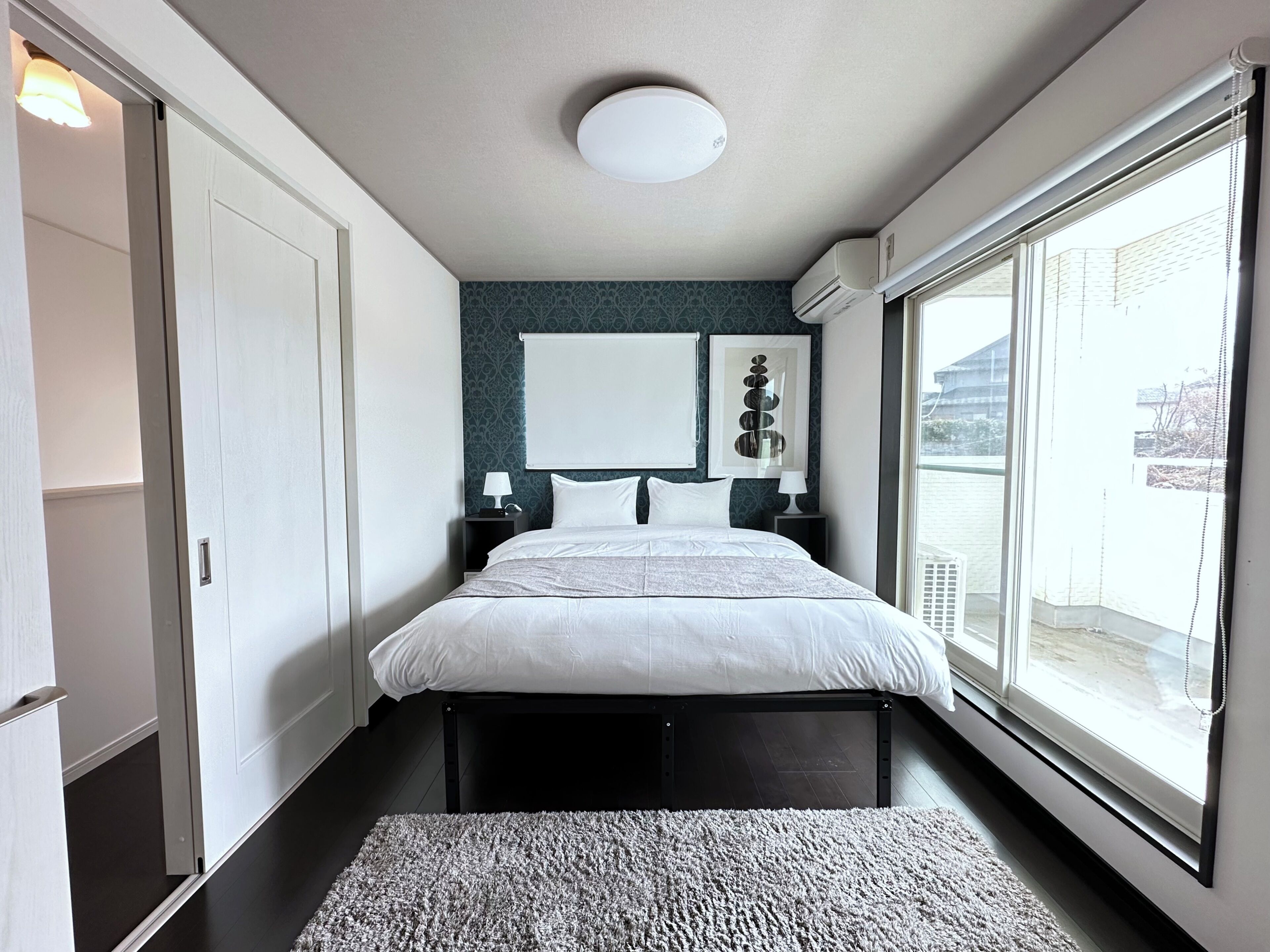 A modern bedroom with a large bed, bedside tables, a wall-mounted air conditioning unit, and a view of the outside through a large window.