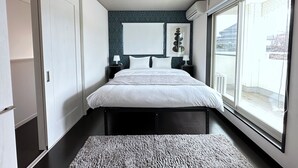 A modern bedroom with a large bed, bedside tables, a wall-mounted air conditioning unit, and a view of the outside through a large window. - Modern Property 15 Mins To Kumamoto Airport By Restavio (Ozu)