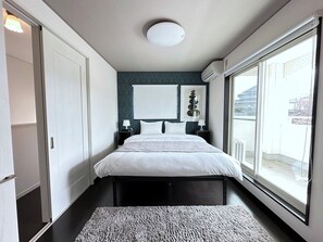 Ett modernt sovrum med en stor säng, sängbord, en väggmonterad luftkonditioneringsanläggning och utsikt över omgivningen genom ett stort fönster.