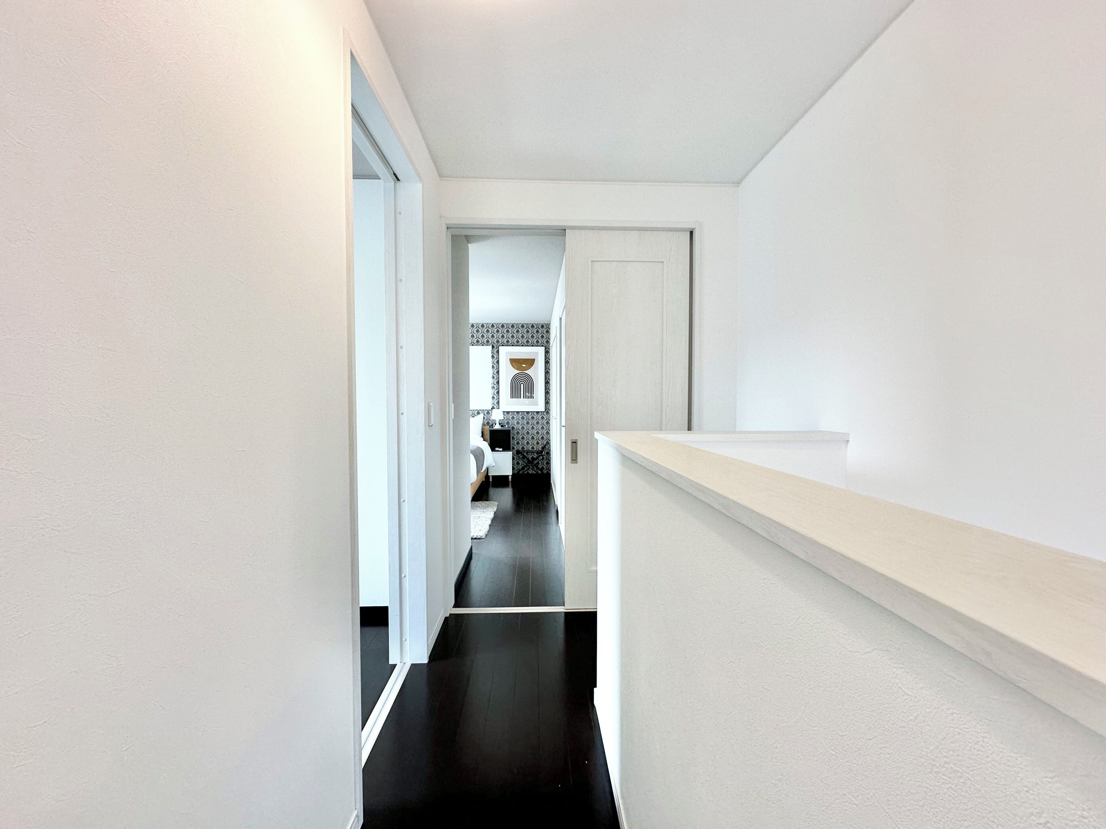 A narrow hallway with a black floor leading to a bedroom with a bed, bedside table, and a wall-mounted artwork.