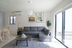 Un salon moderne avec un canapé d’angle gris, une table basse ronde et une grande fenêtre offrant une vue sur l’extérieur.