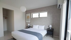 A modern bedroom with a large bed, a bedside table with a lamp, a framed artwork on the wall, and a window providing natural light. - JP Sugimizu Units By Restavio (Ozu)