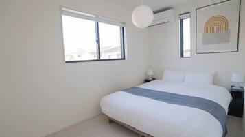 A bedroom with a bed, a bedside table, a lamp, a window, and an air conditioning unit.