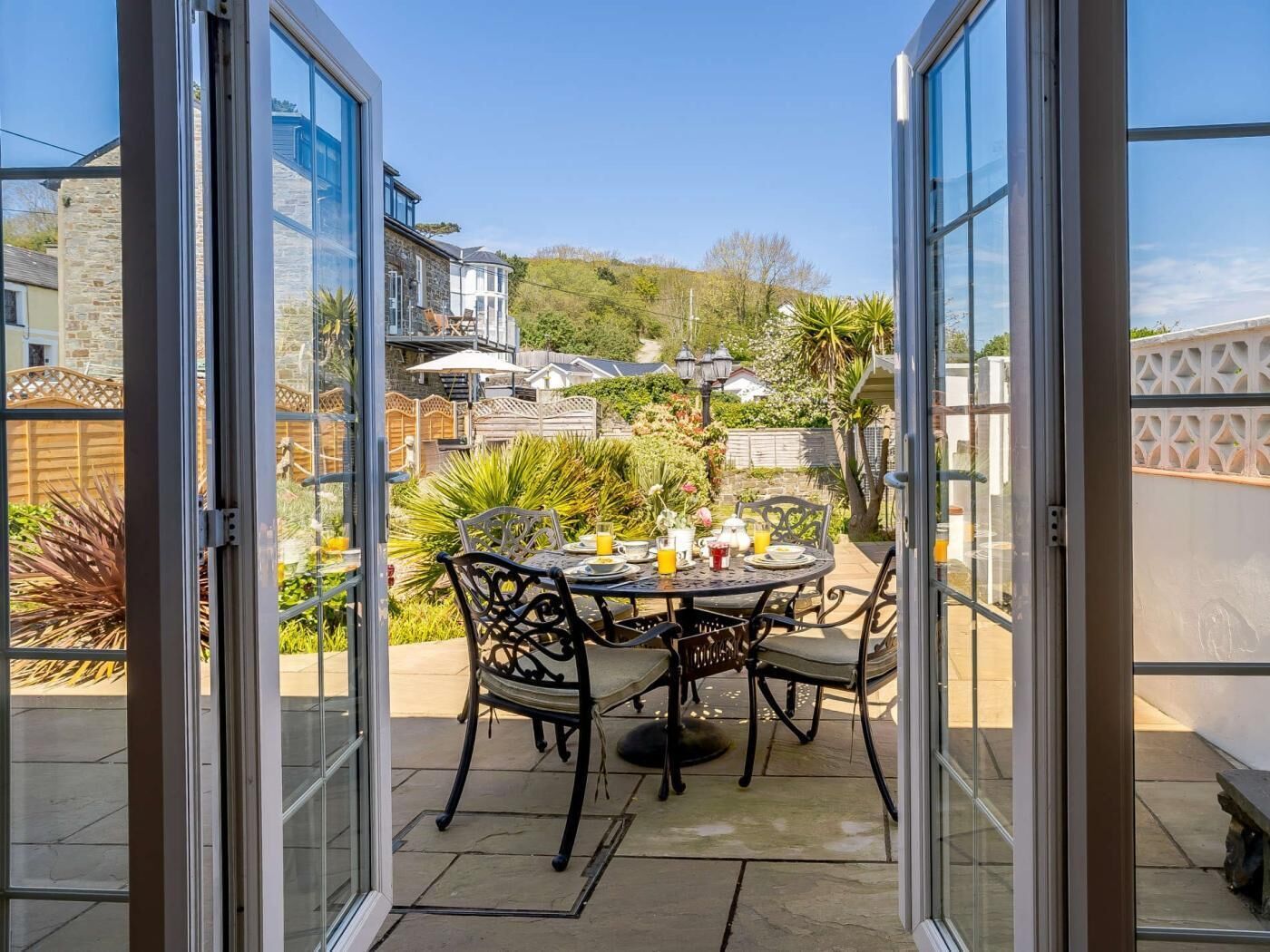 An open patio with a table set for two, surrounded by chairs, and a view of a garden and neighboring houses.