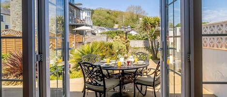 An open patio with a table set for two, surrounded by chairs, and a view of a garden and neighboring houses.