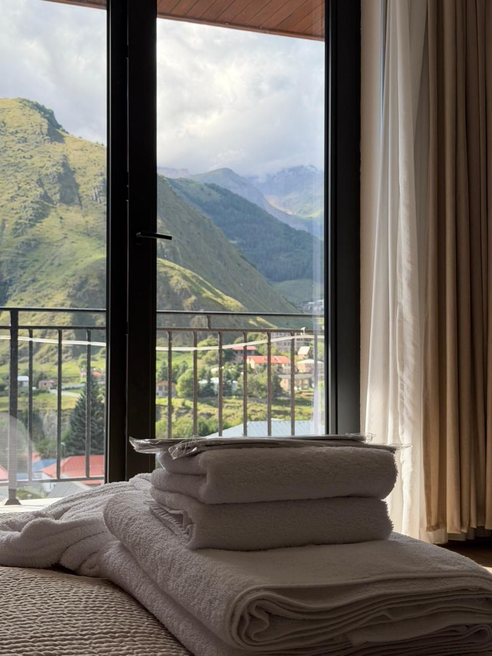 A room with a view of mountains, a balcony with a railing, and a stack of towels on a bed.