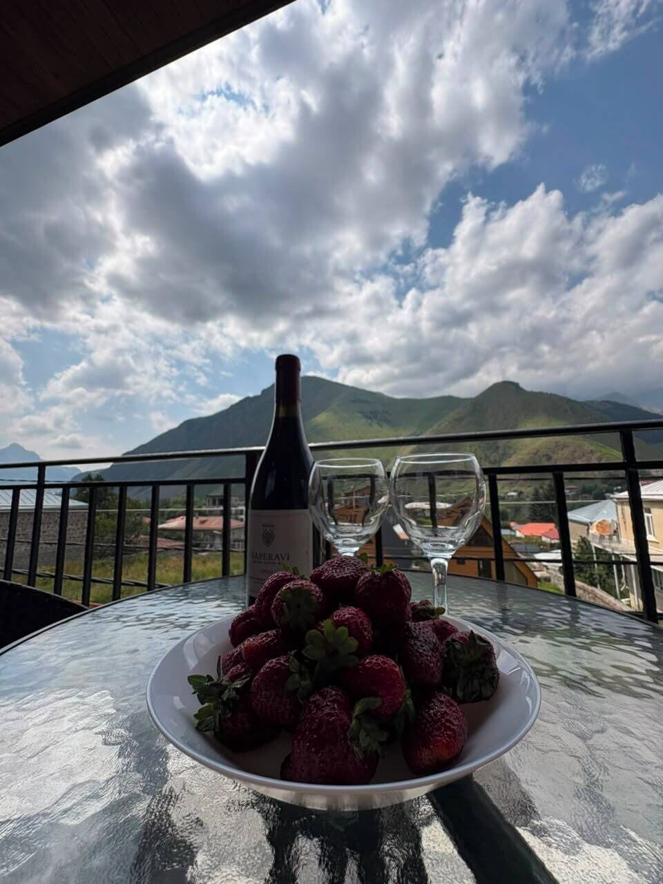 Una mesa con una botella de vino, dos copas y un plato de fresas en un balcón con vista a la montaña.