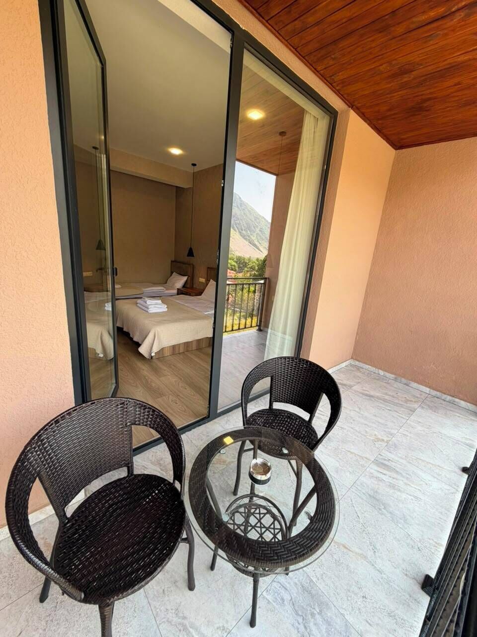 Un balcon avec deux fauteuils en osier et une petite table ronde, une chambre avec deux lits et une vue sur les montagnes, visible à travers une porte vitrée.