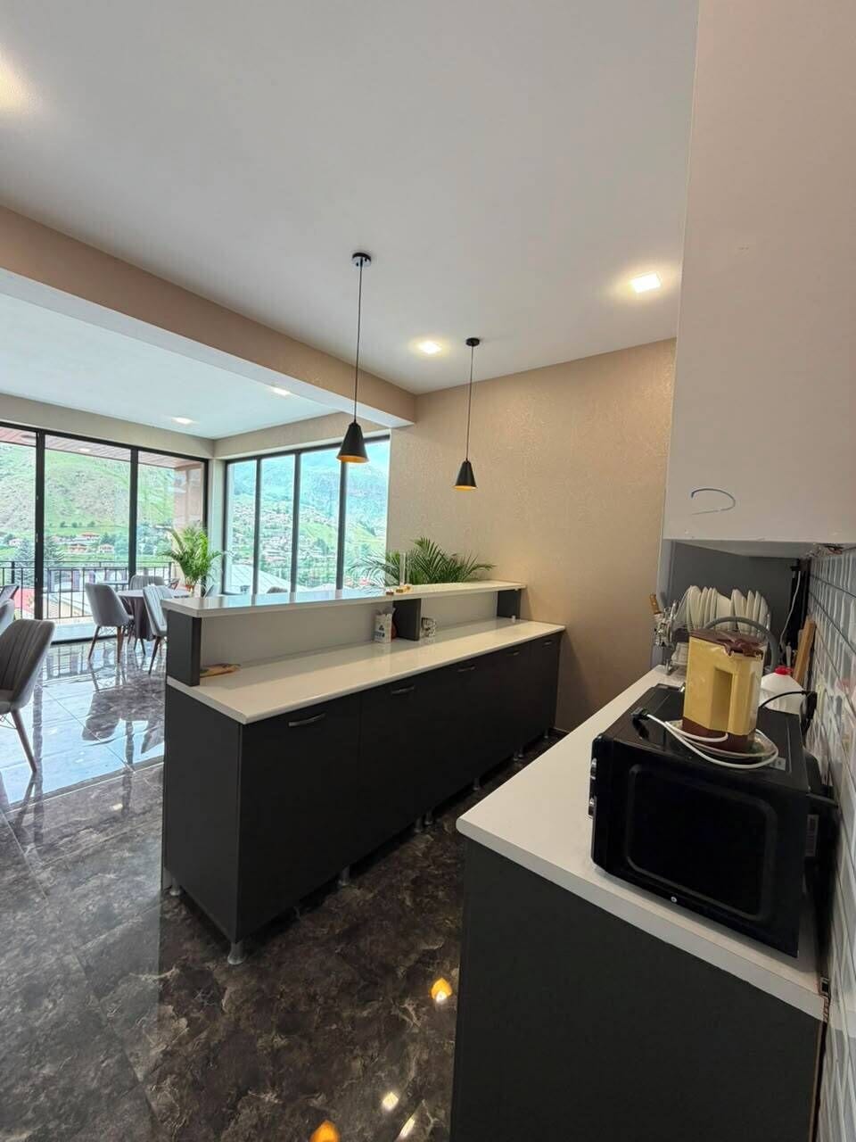 Una cocina moderna con una isla central, un comedor con sillas y vistas a un paisaje verde a través de amplios ventanales.