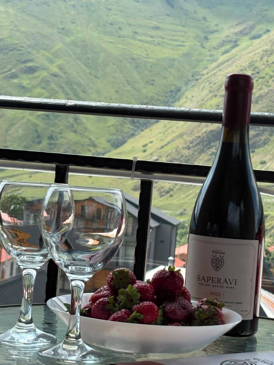 Una botella de vino Saperavi, dos copas de vino vacías y un cuenco con fresas sobre una mesa con vistas a colinas verdes.