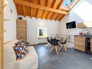 Una habitació acollidora amb un sostre de fusta alt, una taula de dinar amb caires, un sofà amb un coixí colorit i una cuina amb televisió i electrodomèstics.