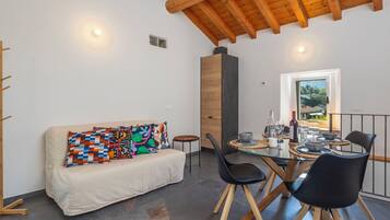Ein modernes Interieur mit Holzbalkendecke, einer Couch mit farbenfrohen Kissen, einem Esstisch mit Stühlen und Blick durch ein Fenster.
