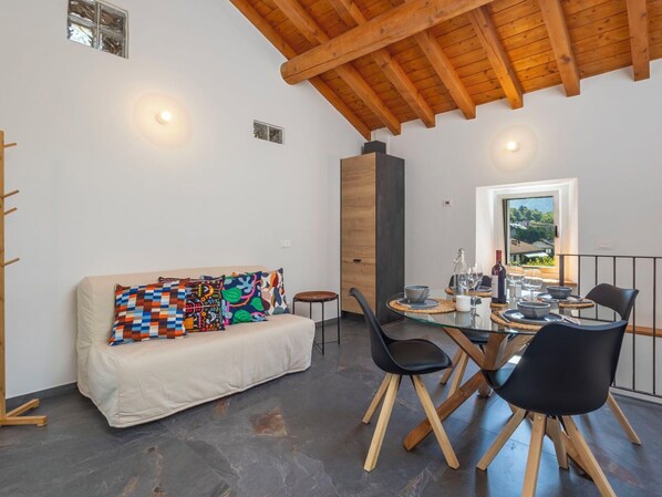 A modern interior with a wooden ceiling, a sofa with colorful cushions, a dining table with chairs, and a view through a window.