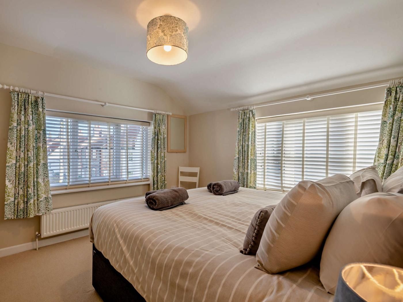 Een slaapkamer met een groot bed, twee ramen met zonwering, een radiator en een hanglamp.