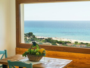 Une table en bois sur laquelle repose un panier en osier rempli de fruits et de légumes, un banc en bois, et une vue sur l’océan et la plage, visible à travers une fenêtre.
