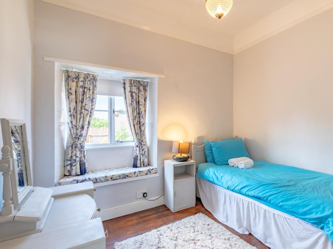 A bedroom with a window, a bedside table with a lamp, a bed with blue bedding, and a white dresser.