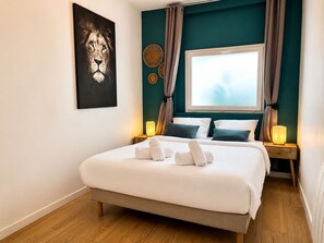Une chambre à coucher avec un lit, deux tables de chevet avec des lampes, une fenêtre avec des rideaux, une œuvre d’art accrochée au mur et un mur d’accent de couleur turquoise.