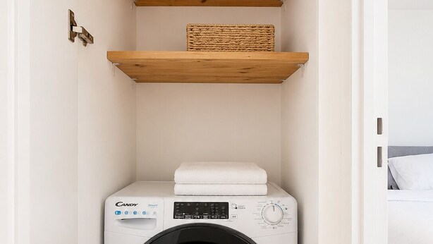 A compact laundry room with a Candy Smart washing machine, wooden shelves, and a bed in the adjacent room. - Le Verdun | Paris & Disneyland (Fontenay-sous-Bois)