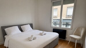 A bedroom with a bed, pillows, a chair, and a window with a view of buildings. - Le Verdun | Paris & Disneyland (Fontenay-sous-Bois)