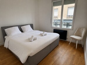Ein Schlafzimmer mit einem Bett, Kissen, einem Sessel und einem Fenster mit Blick auf Gebäude.
