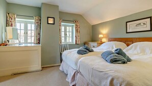 A bedroom with two beds, a window with curtains, a bedside table with a lamp, and a radiator. - 3 Bed in Wells-next-the-sea (oc-ncc68) (Stiffkey)