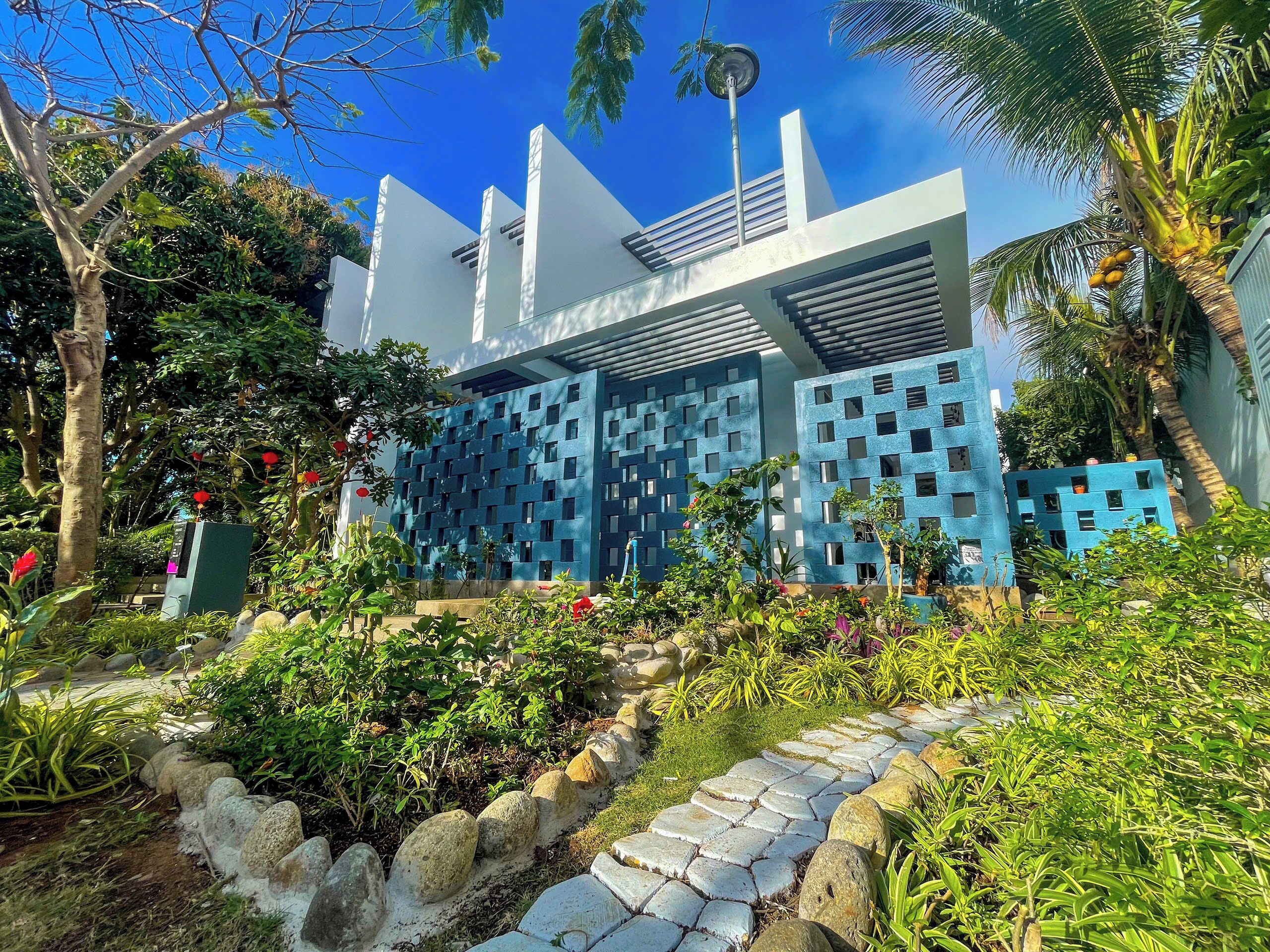 A modern building with a blue facade, surrounded by lush greenery and a stone pathway.